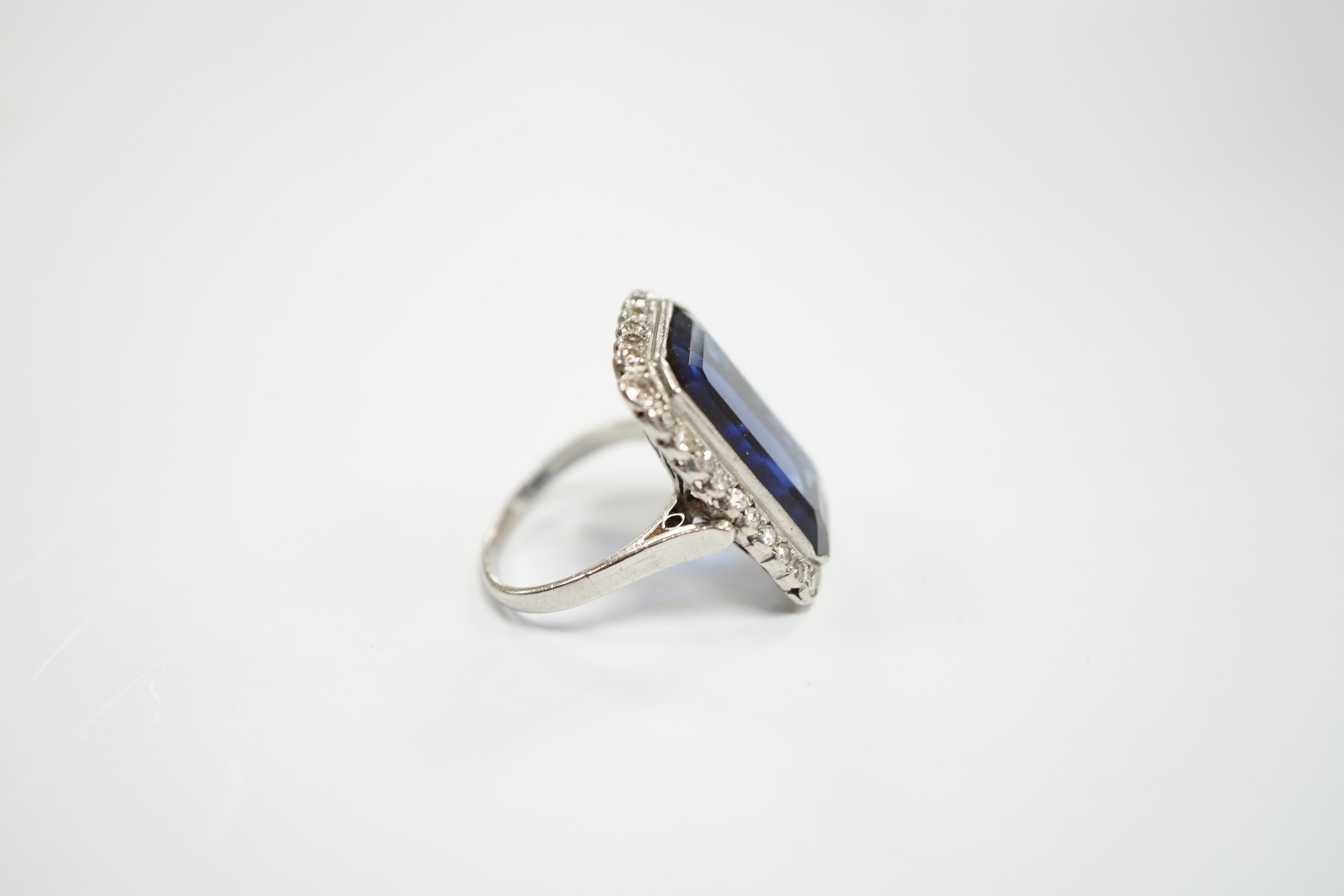 A 1920's white metal and single stone emerald cut synthetic sapphire set dress ring, with millegrain set diamond chip border, size N, gross weight 10 grams.